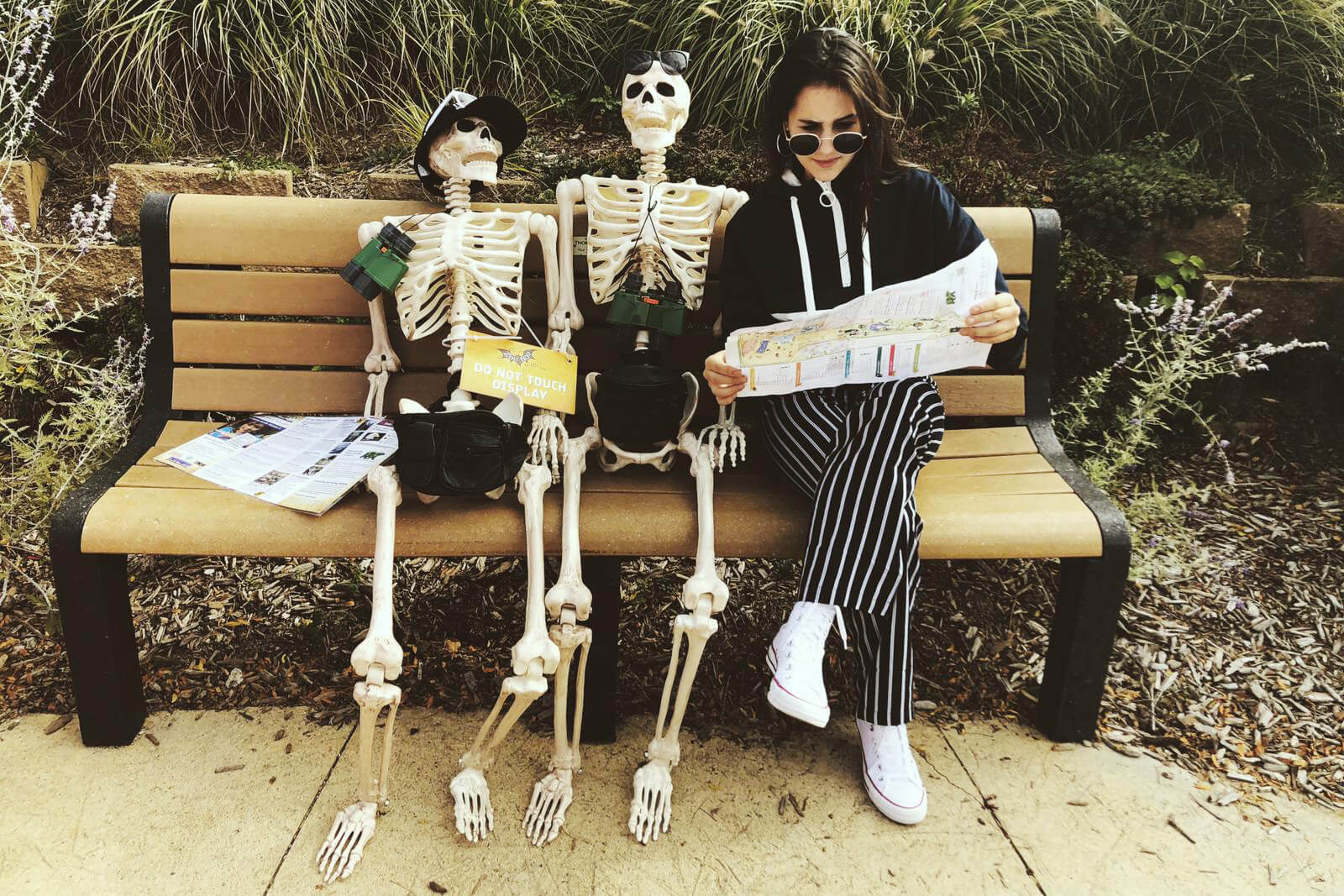 Chica adolescente, sentada en un banco, junto a dos esqueletos, todos personificados como turistas