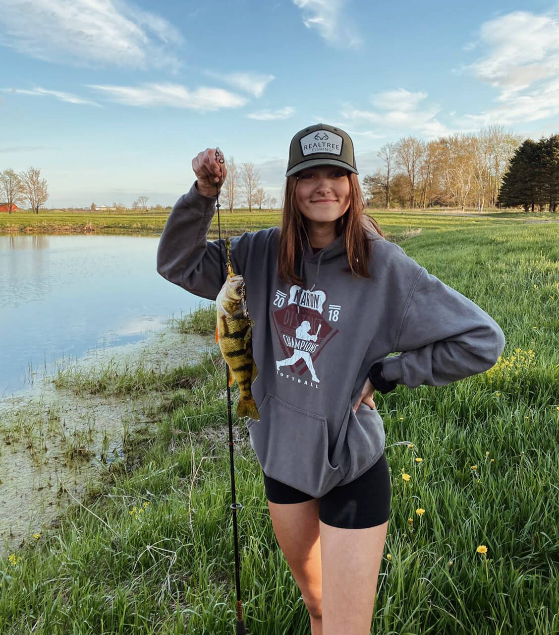 chica joven portando gorra y sudadera, sujetando caña y pescado junto al río