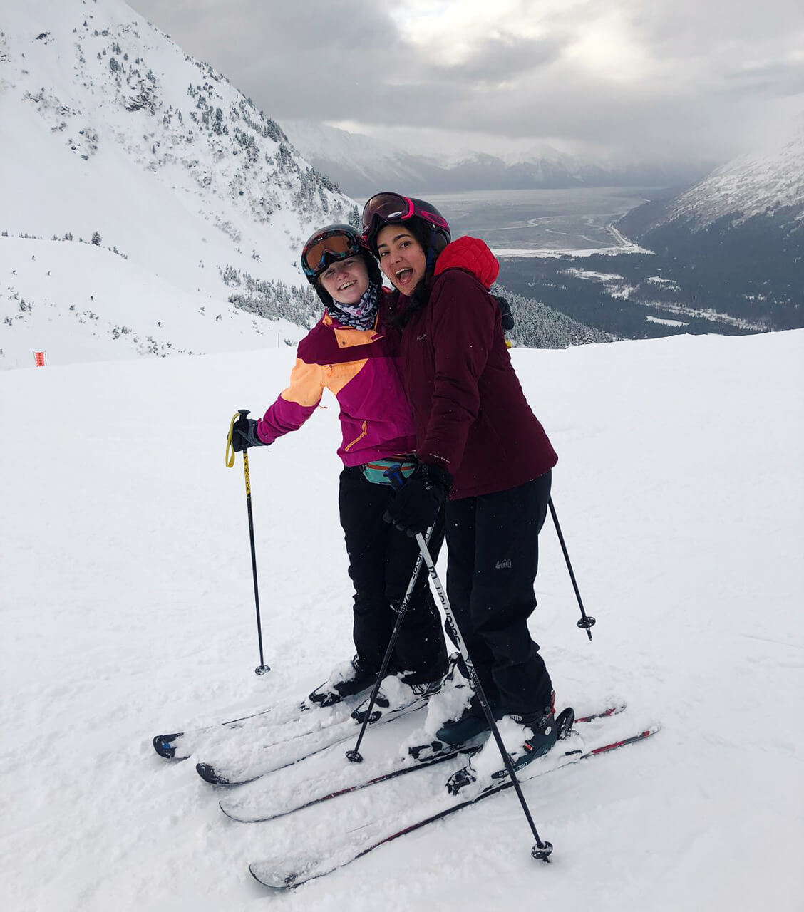 Dos amigas esquiadoras abrazadas en medio de un valle de montañas cubierto en nieve