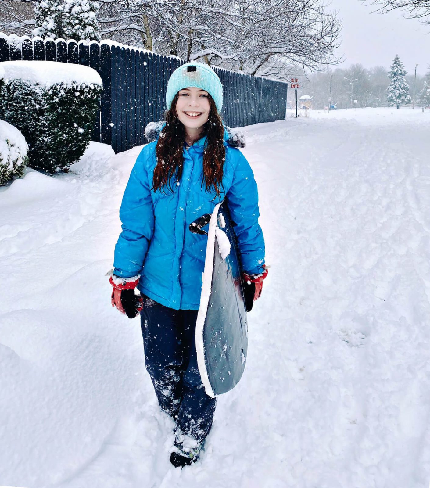 Chica joven y sonriente, bien abrigada y portando tabla, en vecindario cubierto de nieve