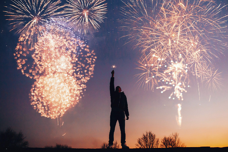 Silueta de una persona celebrando frente a grandes fuegos artificiales bajo un cielo atardecido.