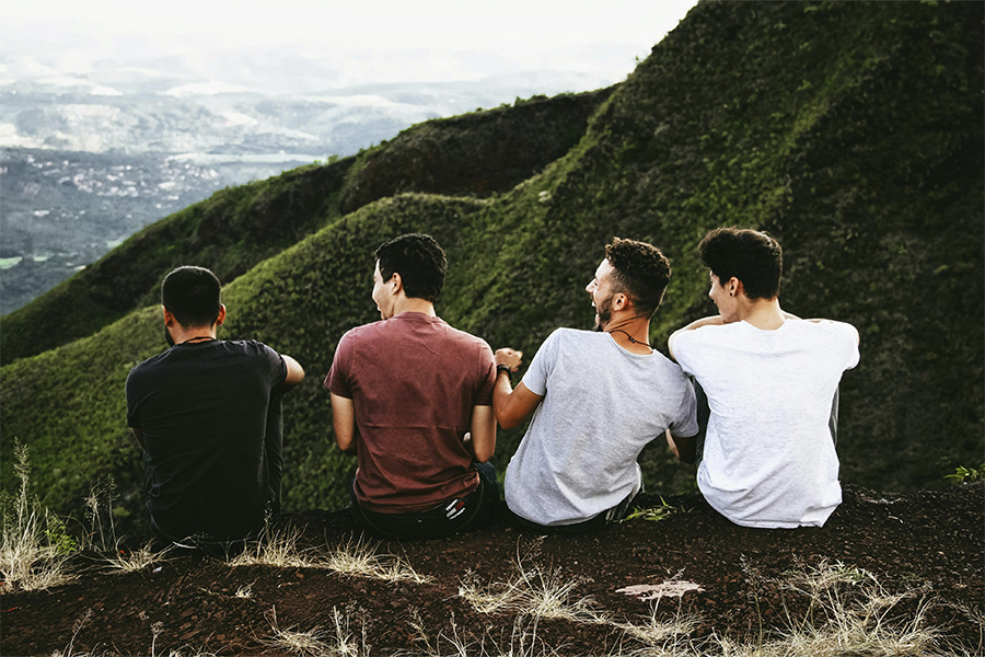 Cuatro amigos jóvenes sentados de espaldas en la cima de una colina verde mirando el paisaje.