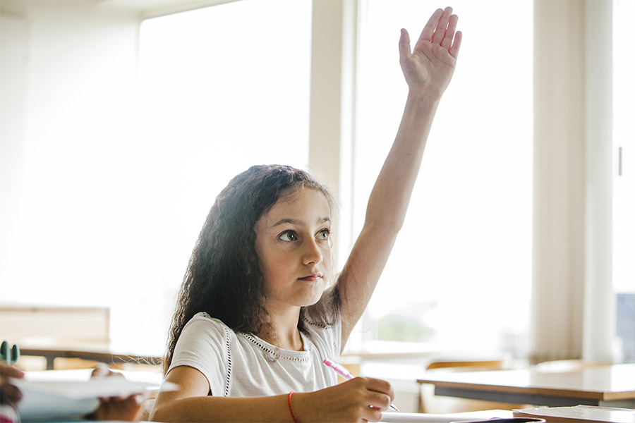 Niña levantando la mano con decisión en un salón de clases para participar en una lección.