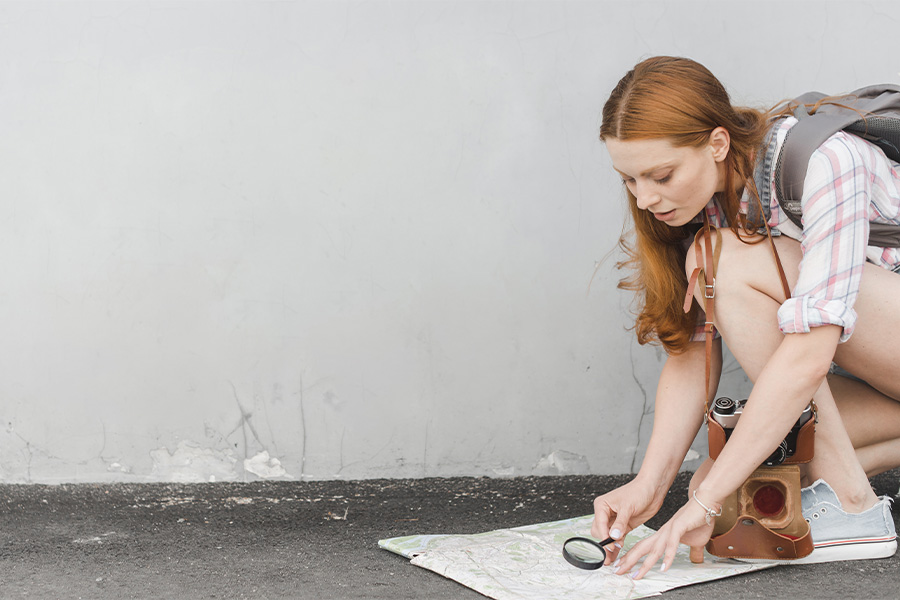 Mujer joven agachada explorando un mapa con una lupa en el suelo durante un viaje de aventura.