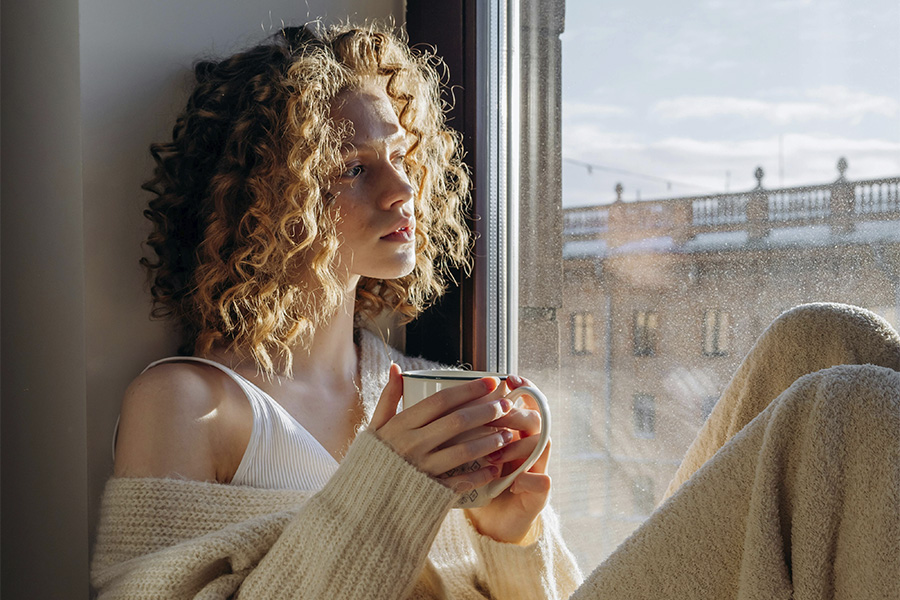 Mujer joven sentada junto a una ventana con una taza en las manos mirando pensativa hacia el exterior.