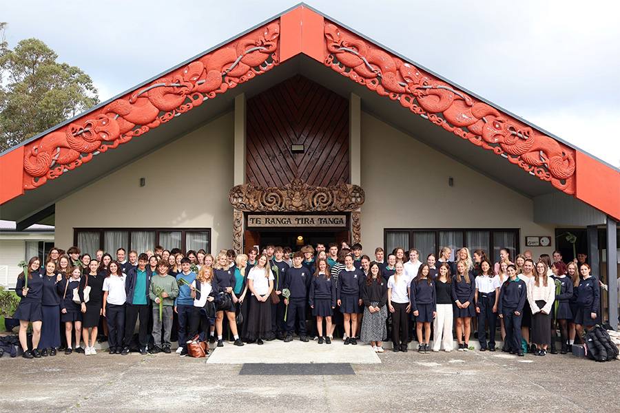 Un gran grupo de estudiantes en uniforme escolar de secundaria posando formalmente frente al wharenui (casa de reuniones) "TE RANGA TIRA TANGA" profusamente tallado en Nueva Zelanda.