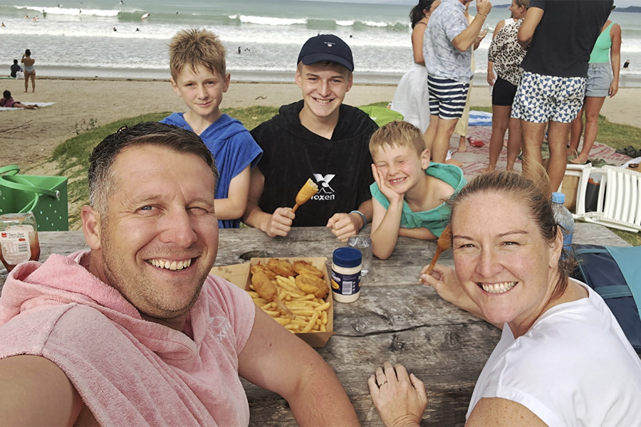 Una familia internacional feliz disfruta de un picnic tradicional de "fish and chips" en una playa de Nueva Zelanda, ilustrando el estilo de vida kiwi que los estudiantes pueden experimentar.