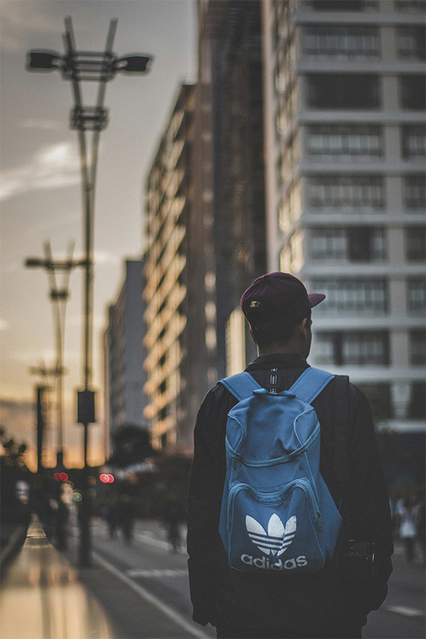 Hombre de espaldas con mochila azul caminando por una calle de grandes edificios urbanos al atardecer.
