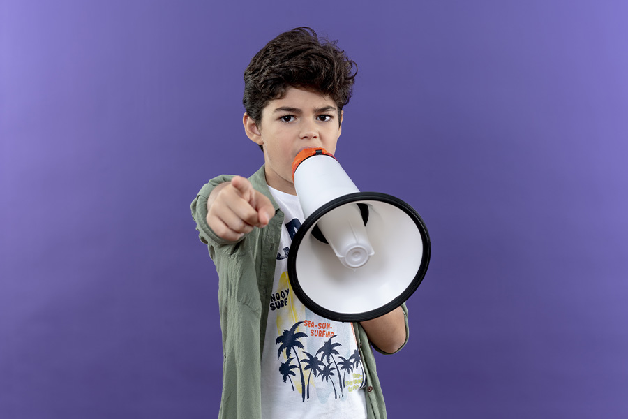 Niño apuntando con el dedo índice hacia el frente mientras sostiene un megáfono blanco y naranja ante un fondo morado.