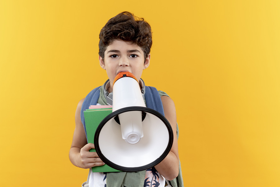 Niño con mochila escolar verde y libros en sus manos sosteniendo un megáfono blanco y naranja frente a un fondo amarillo.