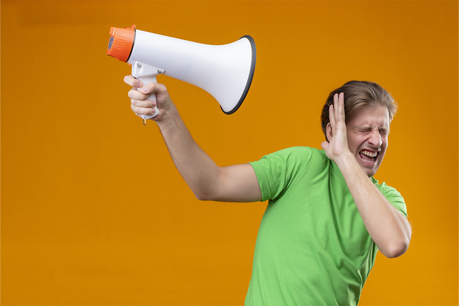Hombre con camiseta verde apartando un megáfono blanco y naranja de su cabeza con expresión de molestia ante un fondo naranja.