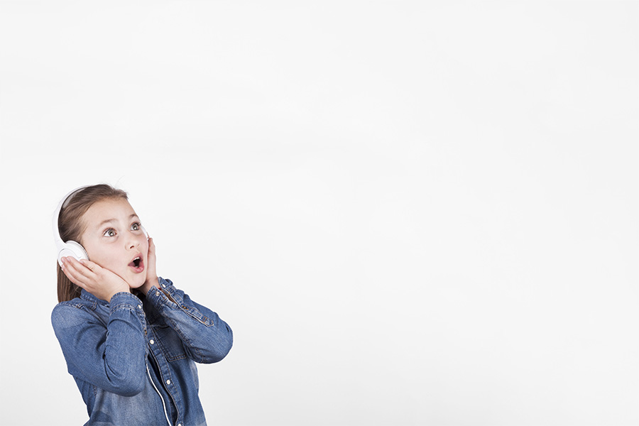 Niña con chaqueta vaquera y auriculares blancos mostrando una expresión de gran asombro con la boca abierta ante un fondo blanco.